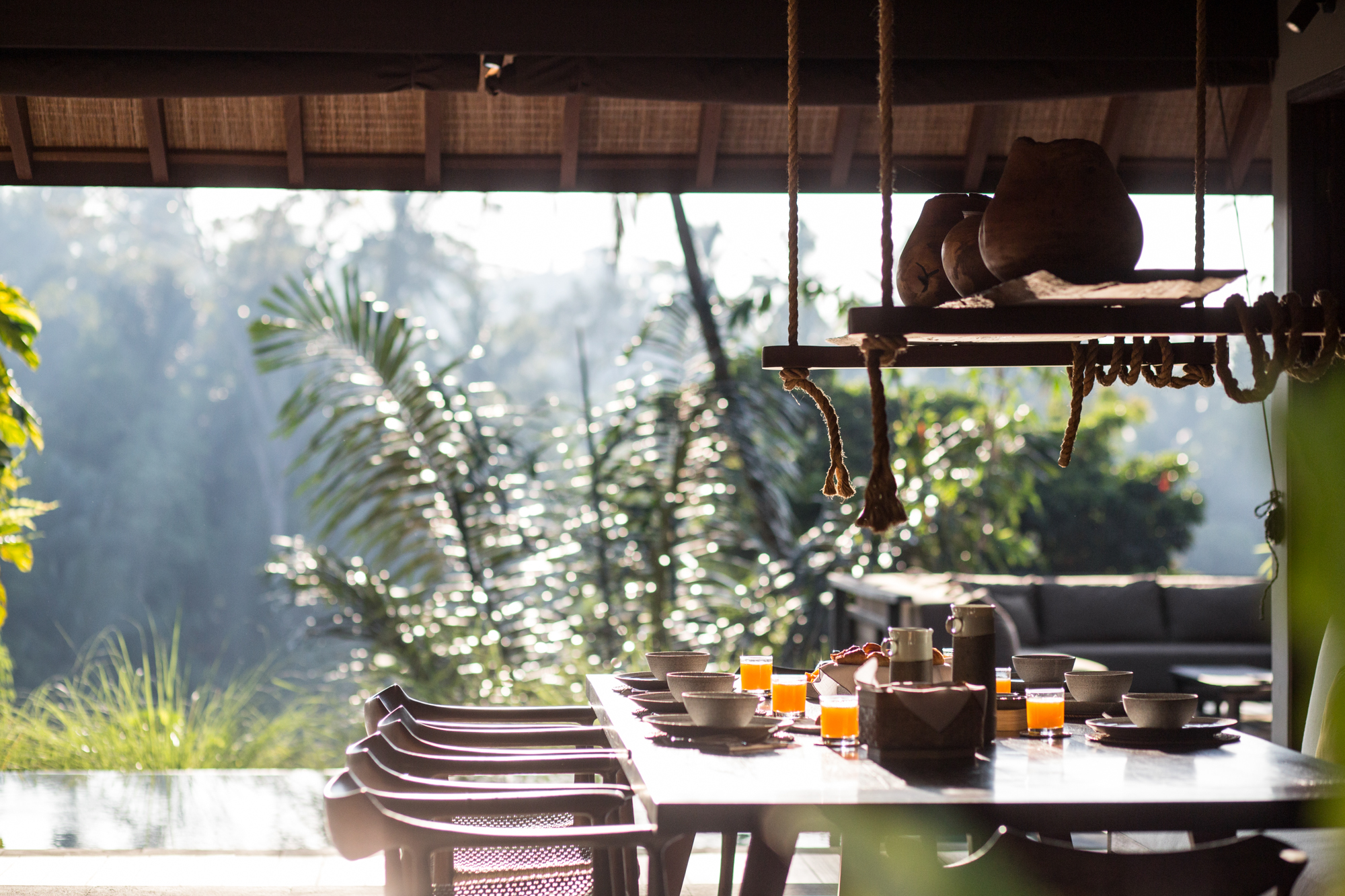 Open-air breakfast table set with ceramic bowls and fresh juice, jungle light beyond