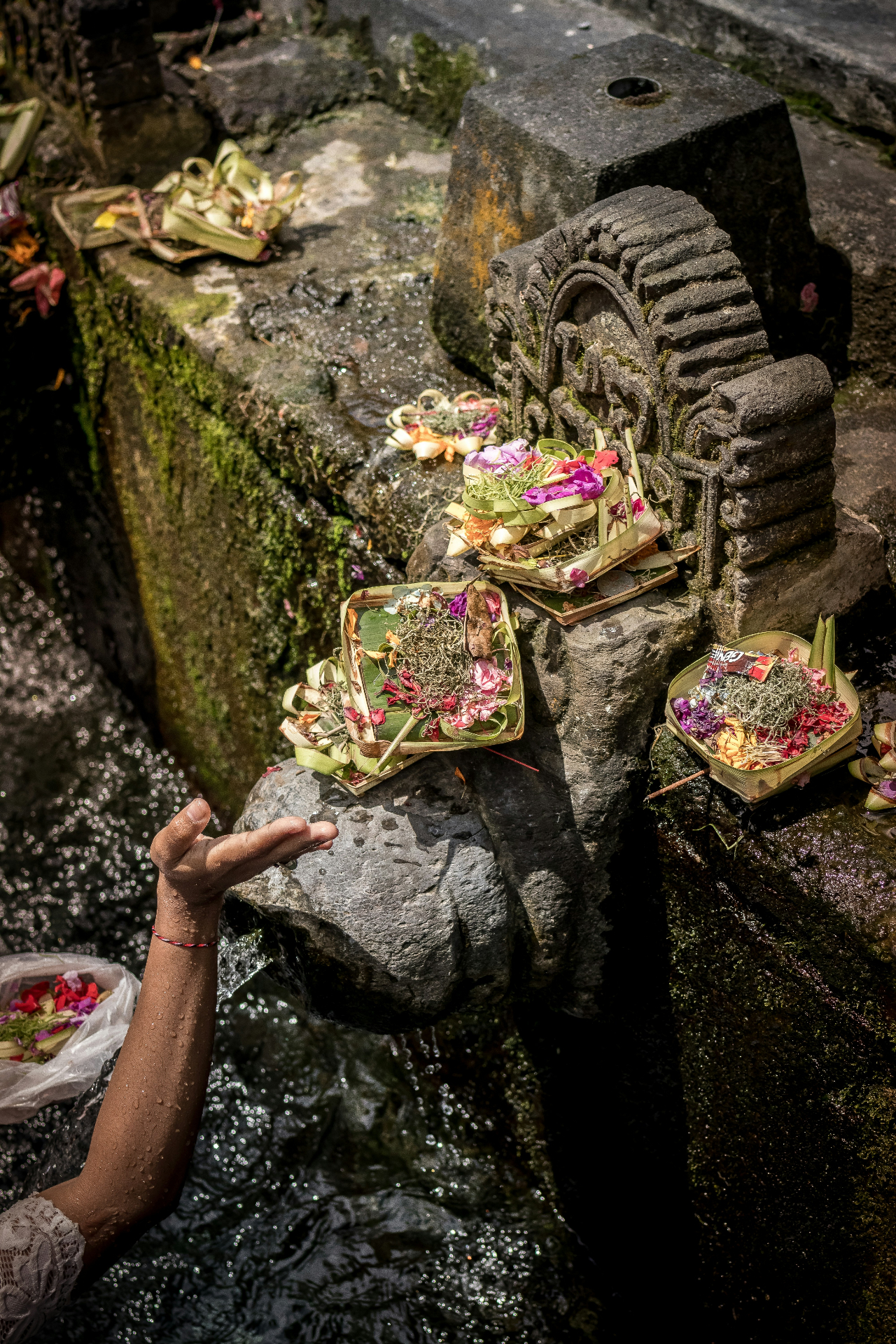An outstretched hand beneath flowing holy water with canang sari flower offerings on a moss-covered stone shrine