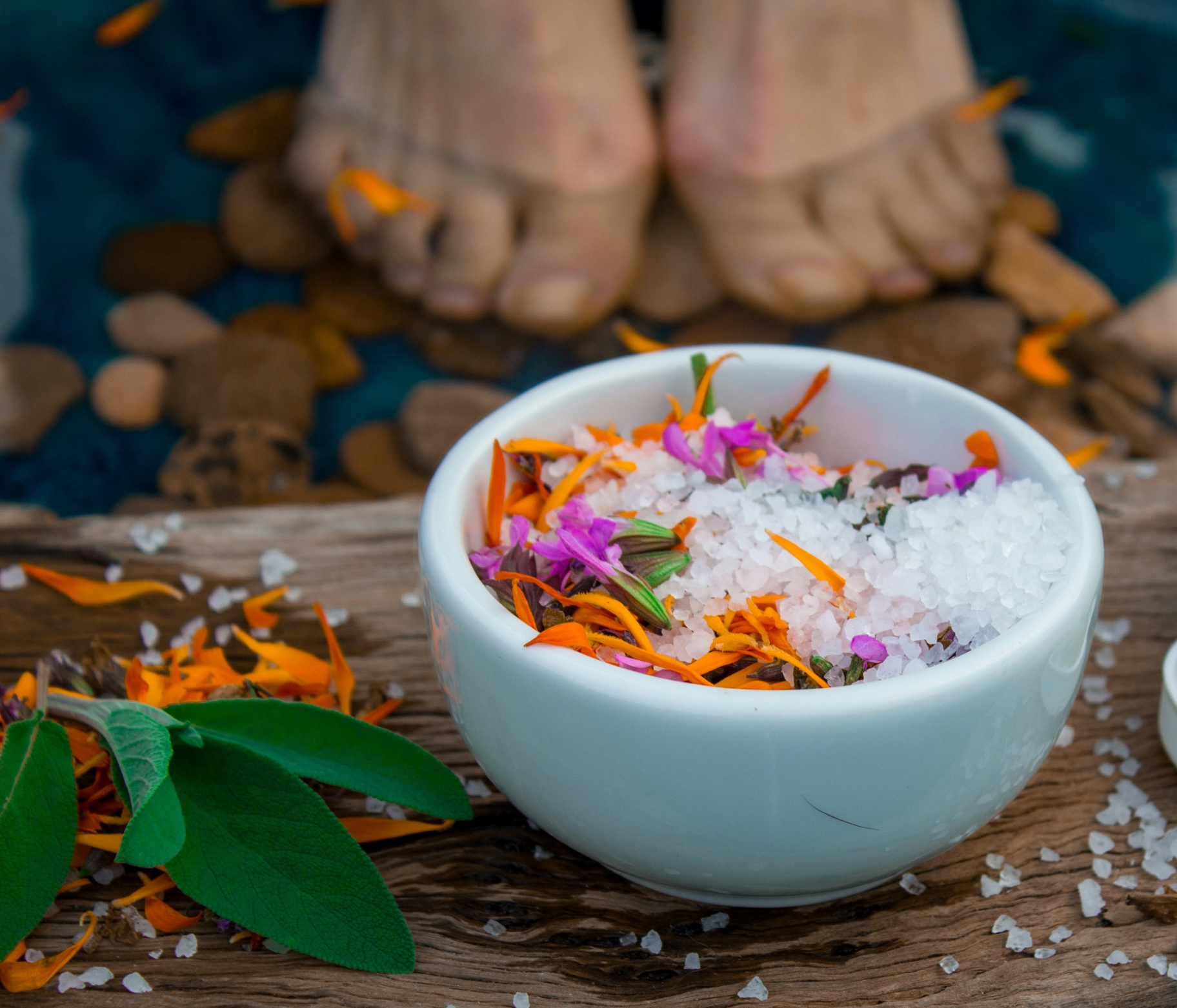Feet resting in water with a bowl of floral salts and sage for the welcome foot scrub